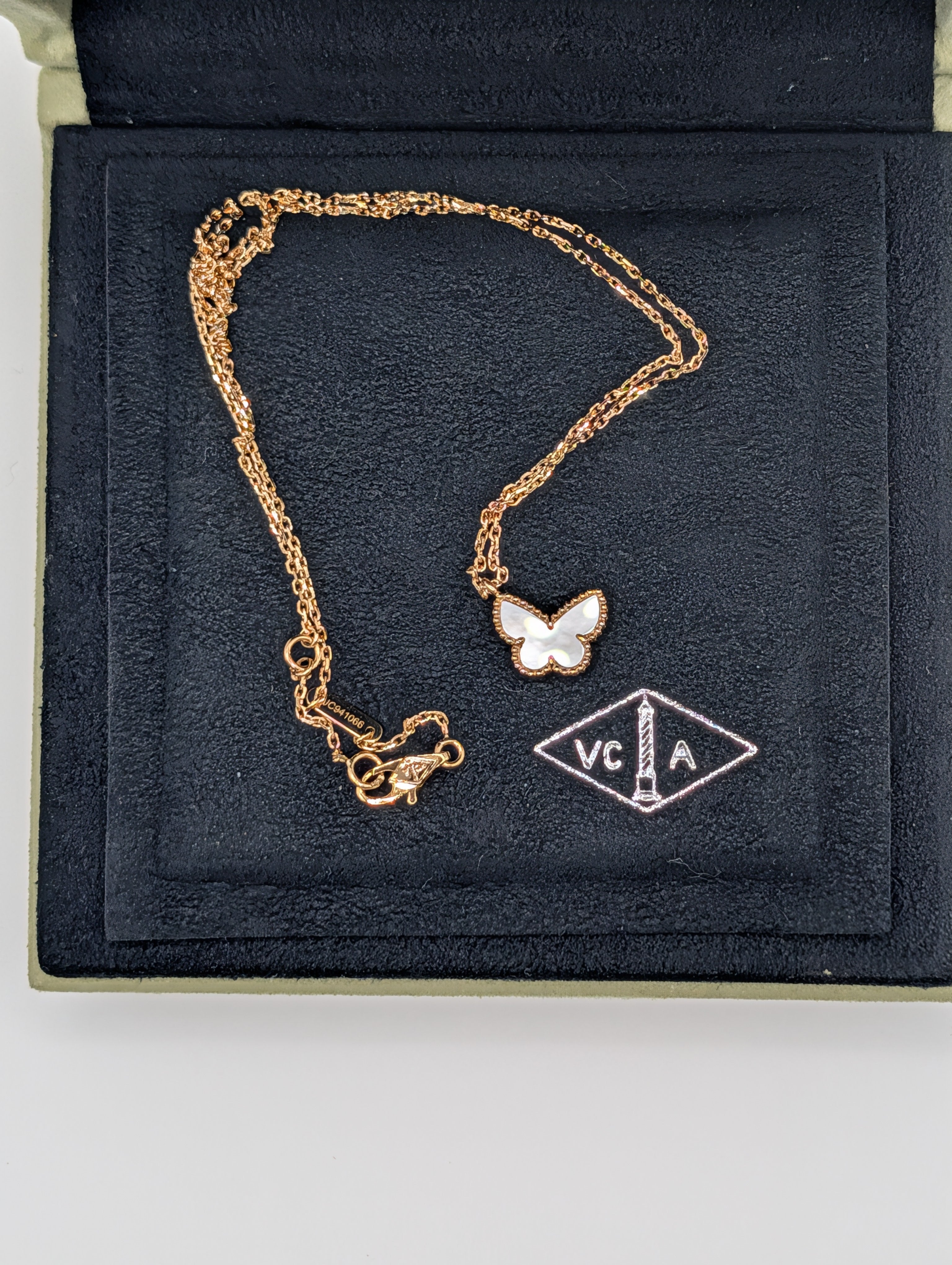 A gold chain necklace with a butterfly pendant featuring white enamel wings, displayed inside a black velvet jewelry box next to a brand tag.